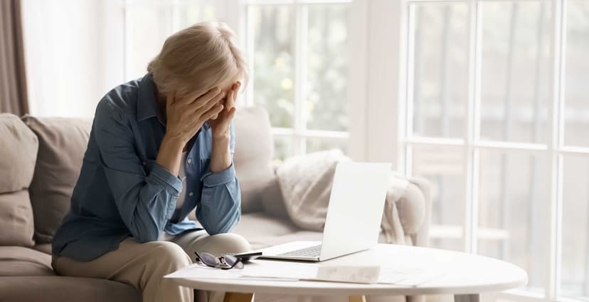 Older woman grieving with her face in her hands while sitting beside a laptop at home, illustrating grief and the use of AI for emotional support.