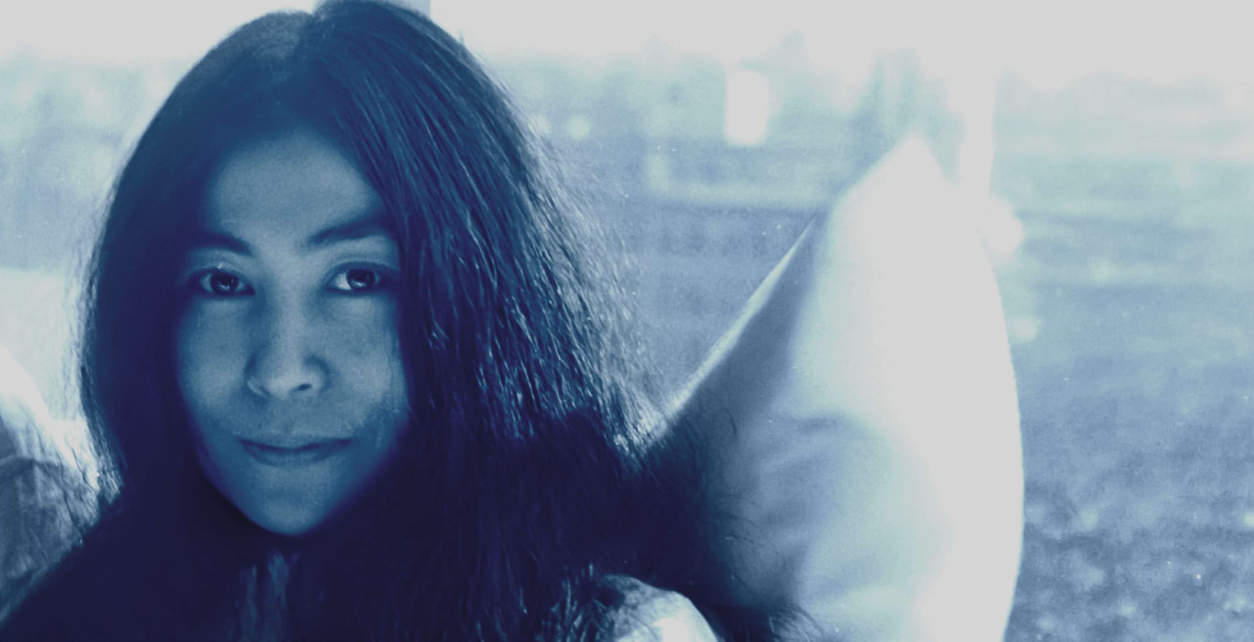 Close-up portrait of Yoko Ono with long dark hair, photographed through a car window in cool blue tones.