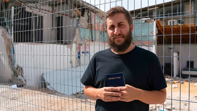 Man holding a Hebrew New Testament in front of a damaged building behind a security fence, reflecting hope and faith amid destruction.