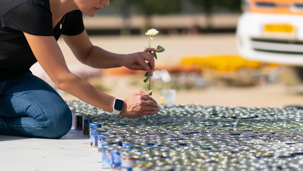 A woman places a small white flower among hundreds of canned goods arranged in rows, creating a moment of beauty amid pain.