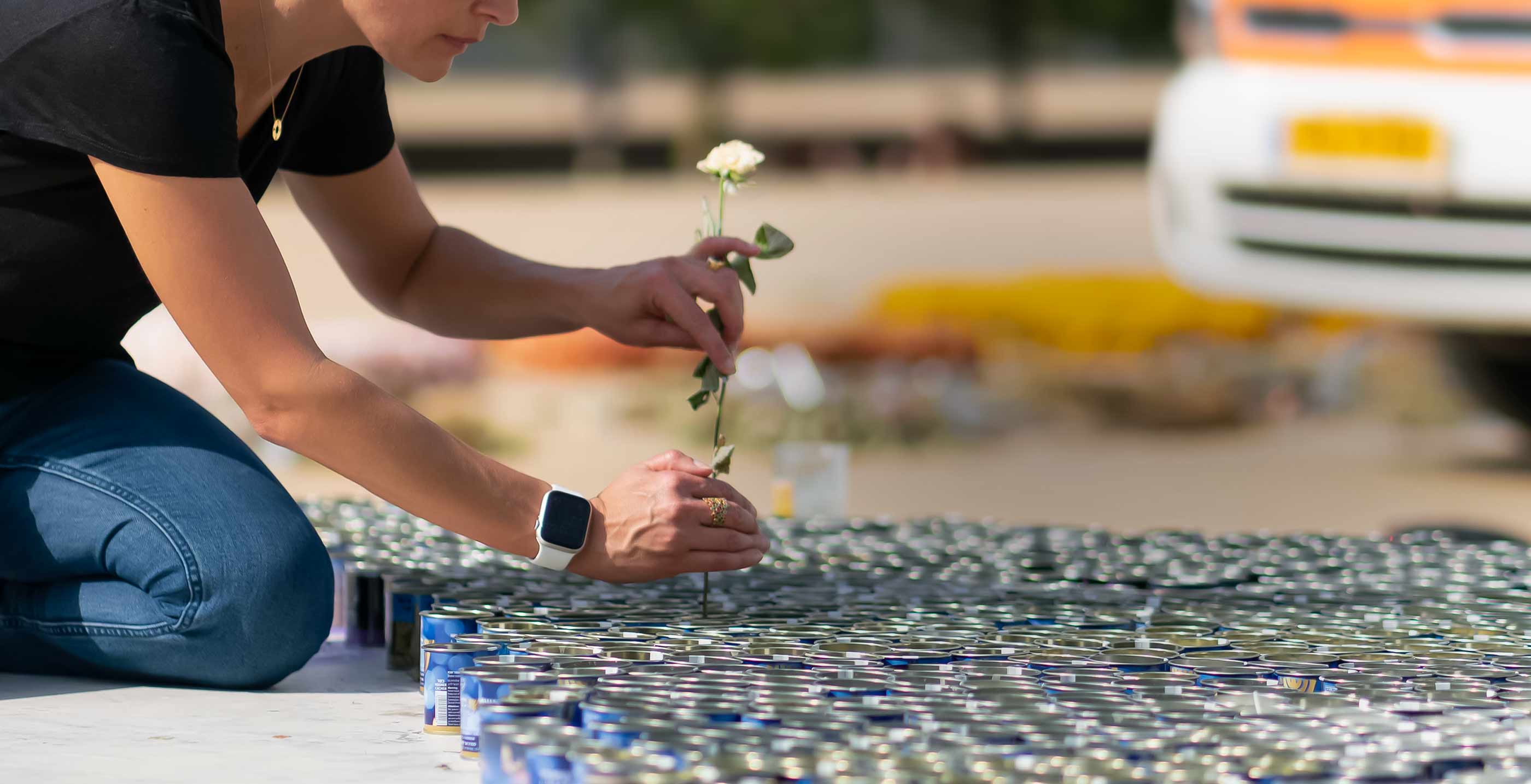 A woman places a small white flower among hundreds of canned goods arranged in rows, creating a moment of beauty amid pain.