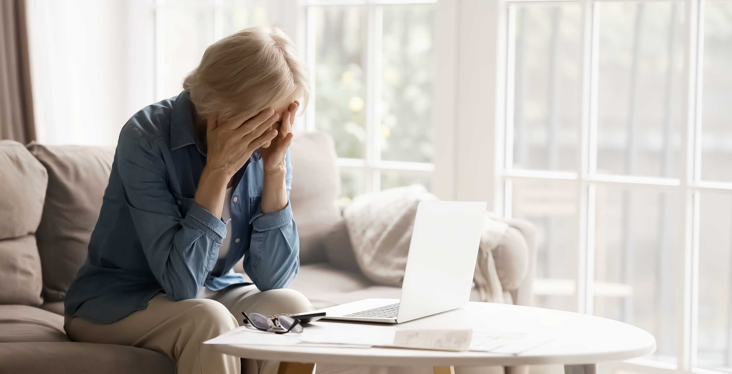 Older woman grieving with her face in her hands while sitting beside a laptop at home, illustrating grief and the use of AI for emotional support.